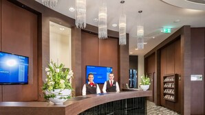 Zona de recepción de hotel con un mostrador curvo, dos recepcionistas, una pantalla digital mostrando un mensaje de bienvenida y una estantería con revistas.