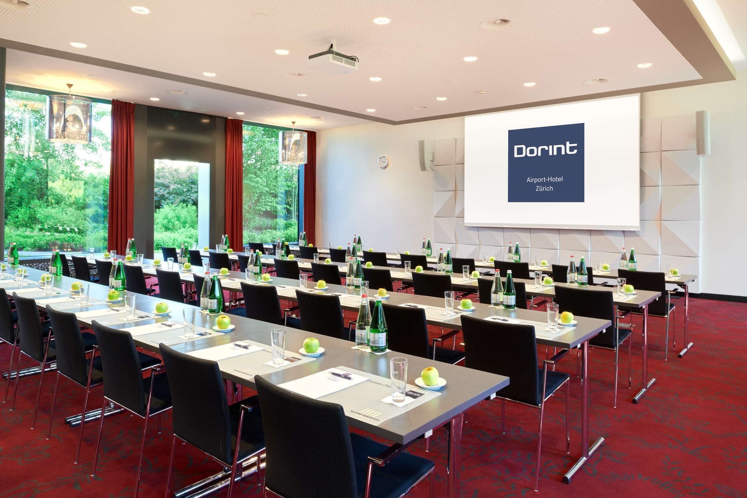 A conference room with rows of tables, chairs, and a projector screen displaying 'Dorint Airport-Hotel Zürich'.