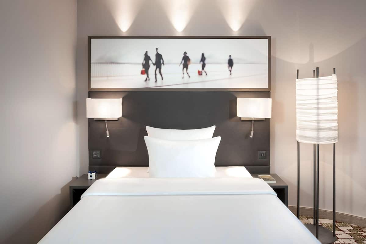 A hotel room with a large bed, two bedside lamps, a flat-screen TV mounted above the bed, and a floor lamp beside the bed.