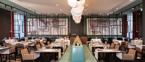 Un restaurante con un largo banco color aguamarina, mesas con manteles blancos y un candelabro moderno.