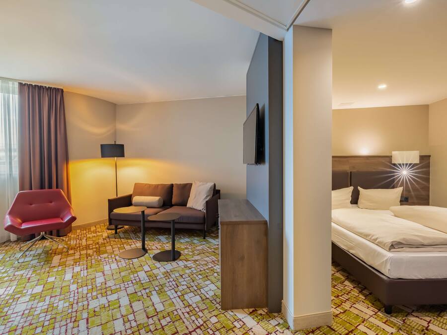 A hotel room with a bed, a sofa, a red chair, a small table, and a TV.