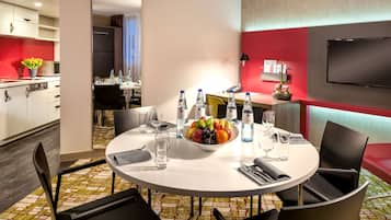 Un'area ristorazione di un hotel con un tavolo rotondo apparecchiato per quattro persone, una ciotola di frutta, bottiglie d'acqua e una TV a schermo piatto montata a parete.