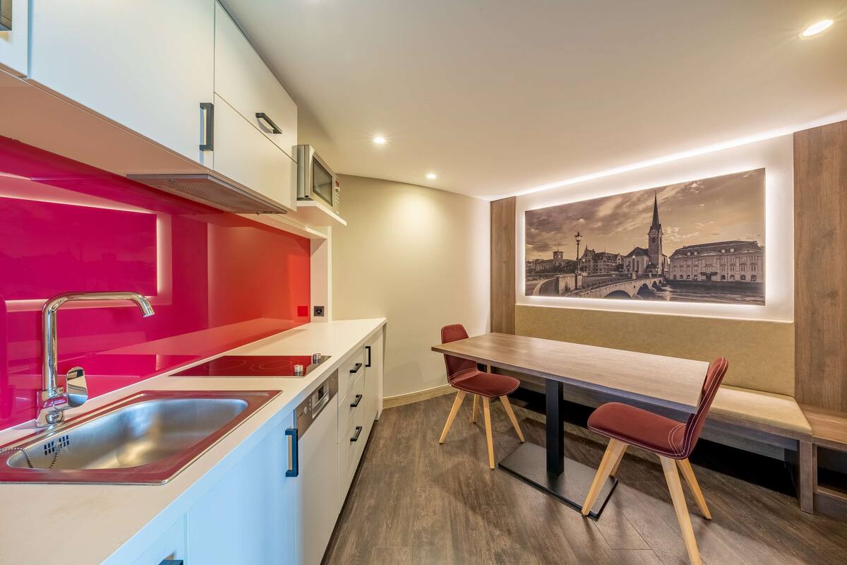 A modern kitchen with a red accent wall, stainless steel sink, and a dining area with a wooden table and red chairs.