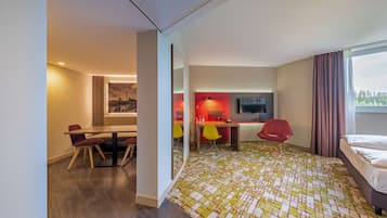 A modern hotel room with a bed, a desk with a chair, a small table, and a large window with curtains.