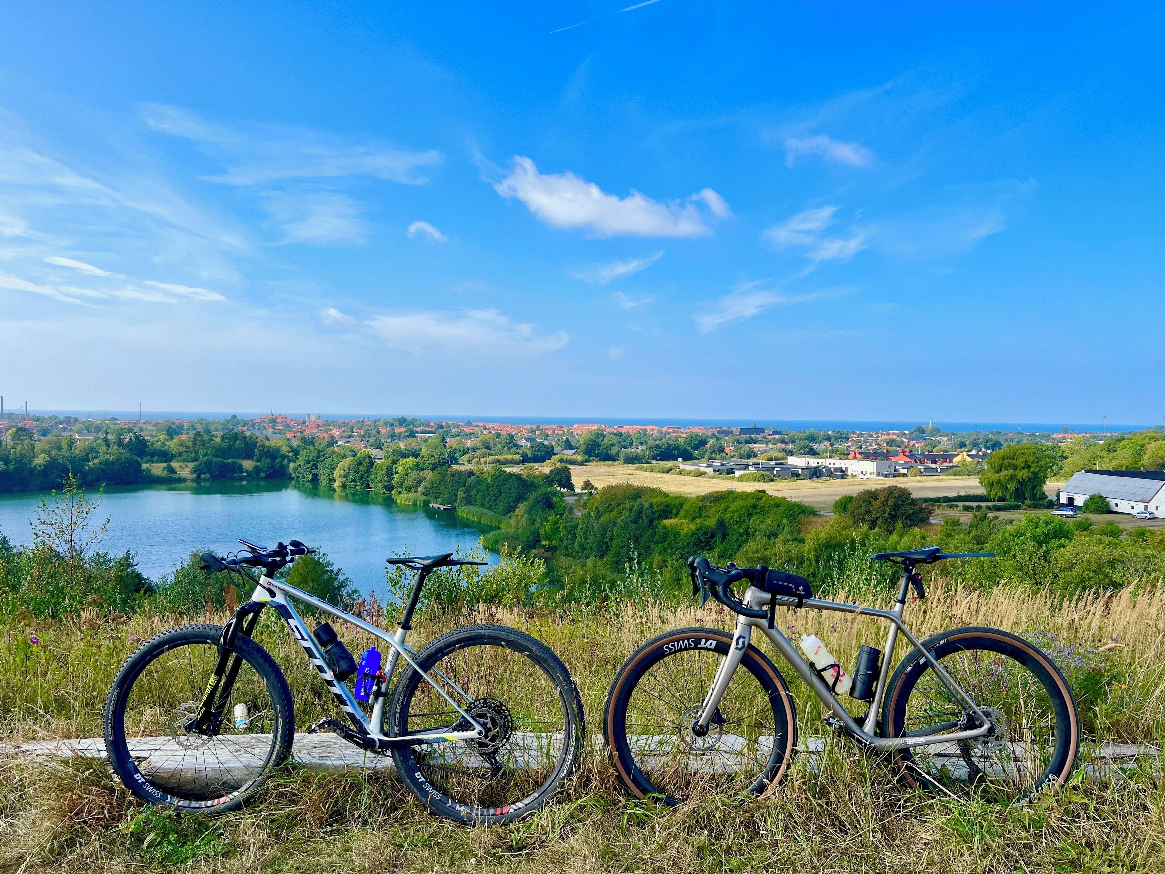 Two bicycles parked on a grassy hill overlooking a lake and a town.