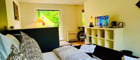 A bedroom with a bed, pillows, a desk with a lamp, a television, and a window with a view of greenery.