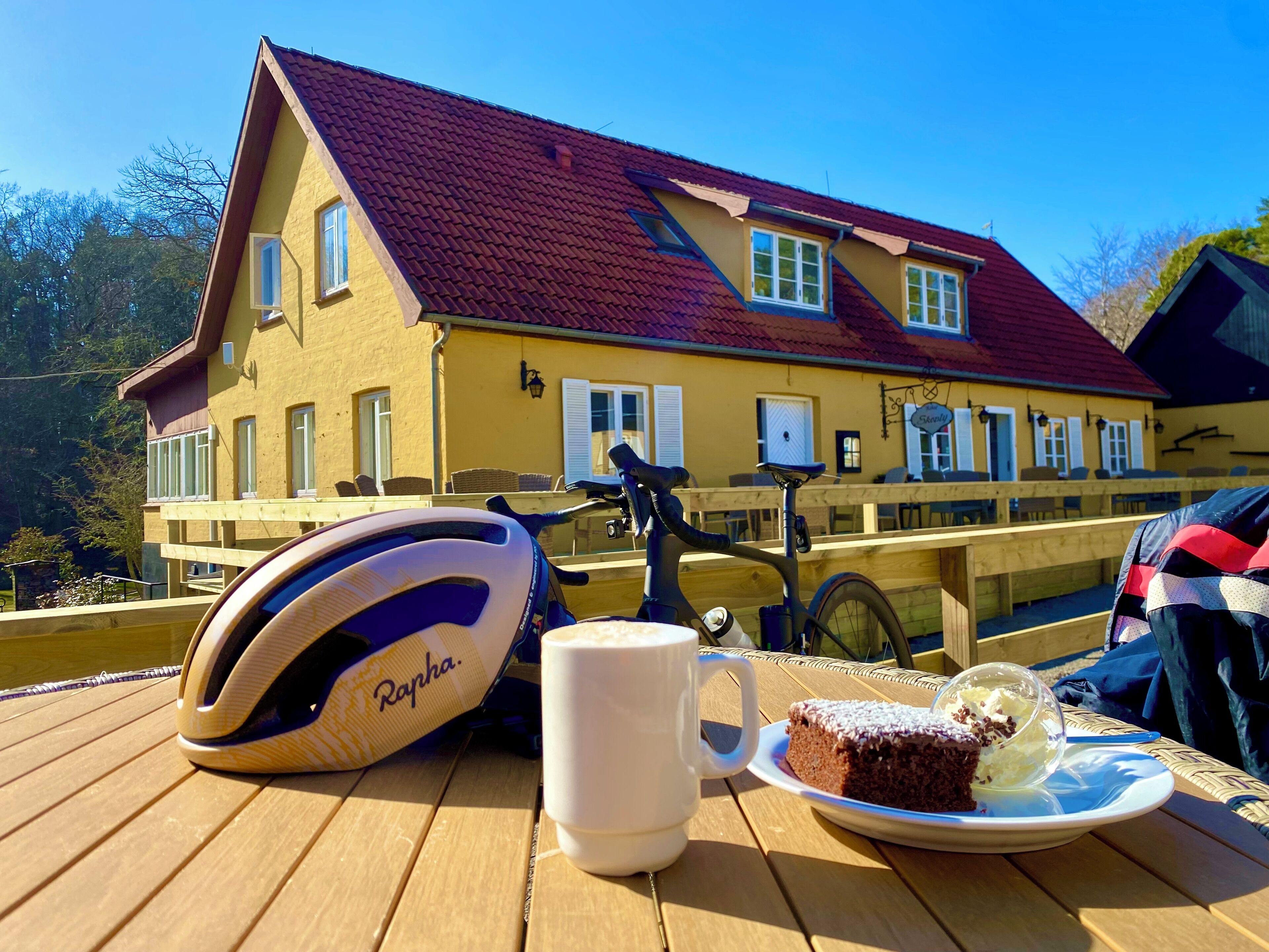 A bicycle helmet, a mug, and a slice of cake on a wooden table with a view of a yellow building with a red roof.