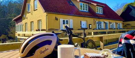 A bicycle helmet, a mug, and a slice of cake on a wooden table with a view of a yellow building with a red roof.