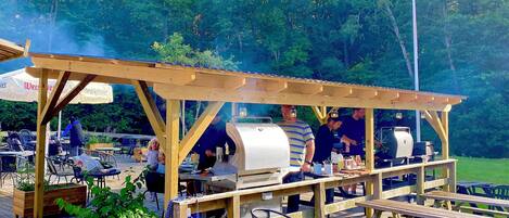 An outdoor wooden structure with a barbecue area, tables, and chairs, surrounded by greenery.