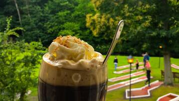 Un dessert servi dans un verre, posé sur une assiette blanche, avec une cuillère à l’intérieur. En arrière-plan, on aperçoit des arbres et un parc avec un chemin rouge et blanc.