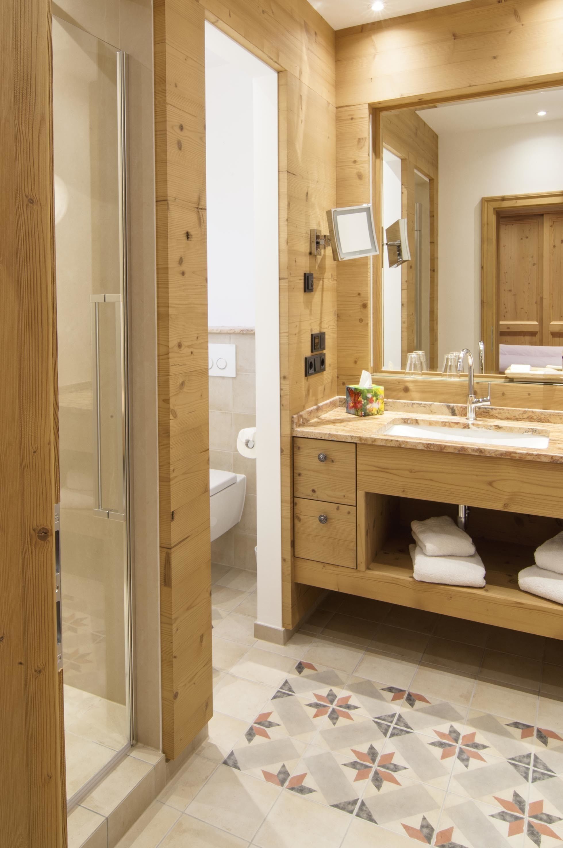 A modern bathroom with wooden paneling, a glass shower enclosure, a sink with a mirror, and a patterned tile floor.
