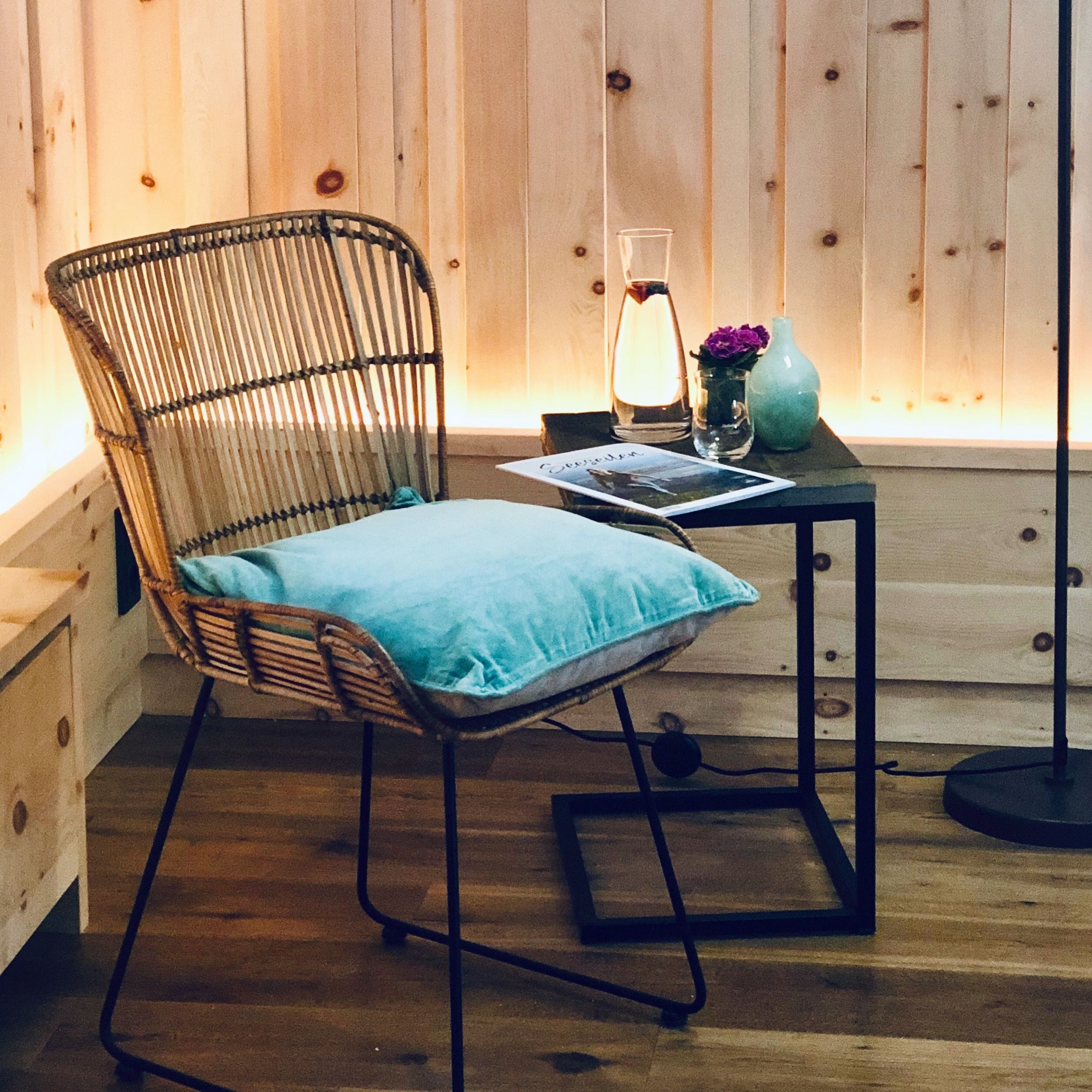A wicker chair with a blue cushion, a table with a glass vase and a candle, and a wooden wall in the background.