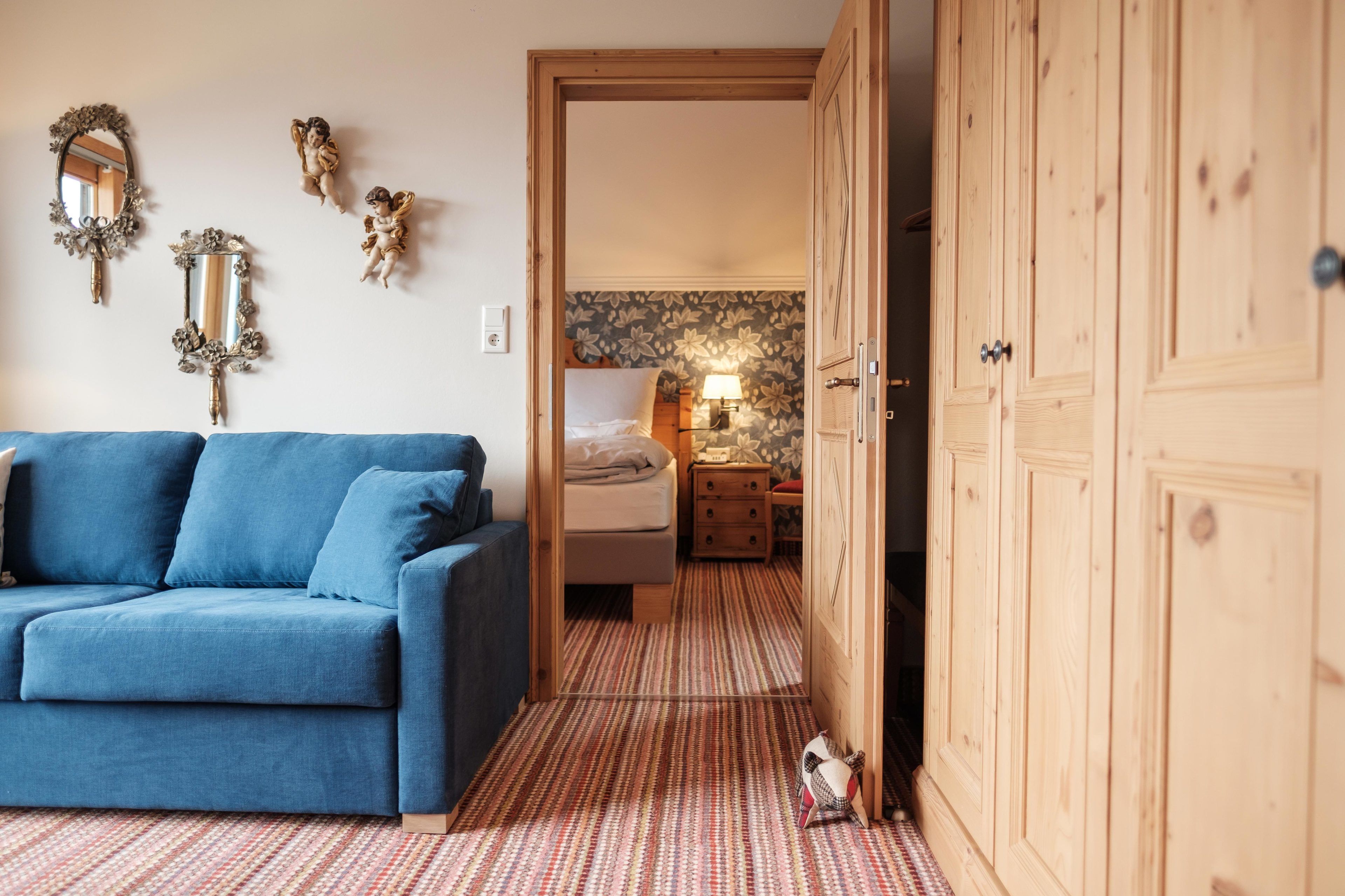 A room with a blue sofa, a wooden wardrobe, and a bed with a floral wallpaper.