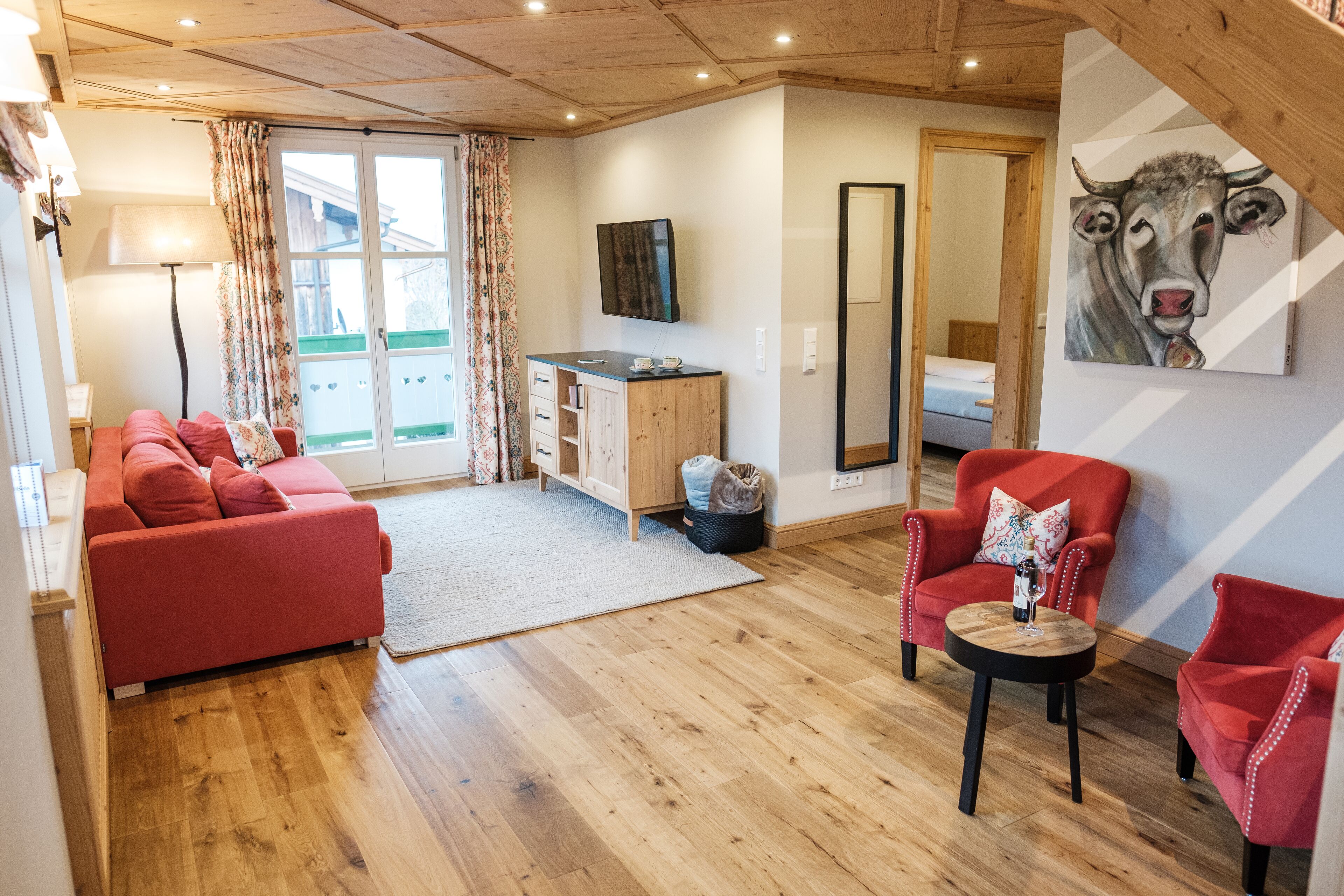 A cozy living room with a red sofa, a wooden cabinet, a small table, and a painting of a cow on the wall.