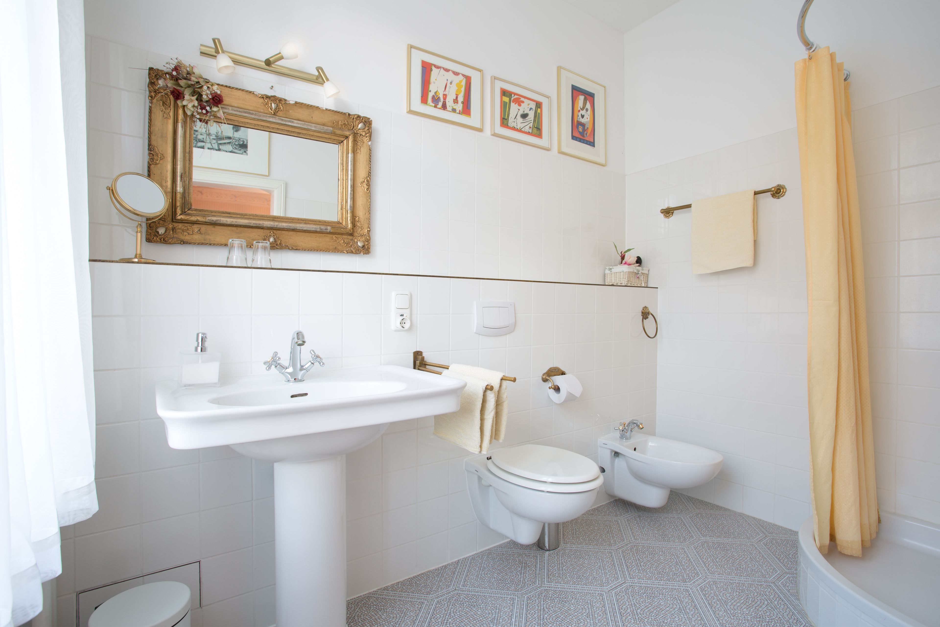 A modern bathroom with a pedestal sink, toilet, bidet, and bathtub. There are framed pictures on the wall and a yellow shower curtain.