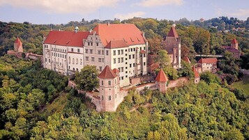 Um edifício histórico imponente com várias torres e um telhado vermelho, rodeado por vegetação exuberante.