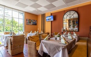 Restaurante com cadeiras e mesas de vime, uma televisão na parede e uma grande janela com vista para vegetação.
