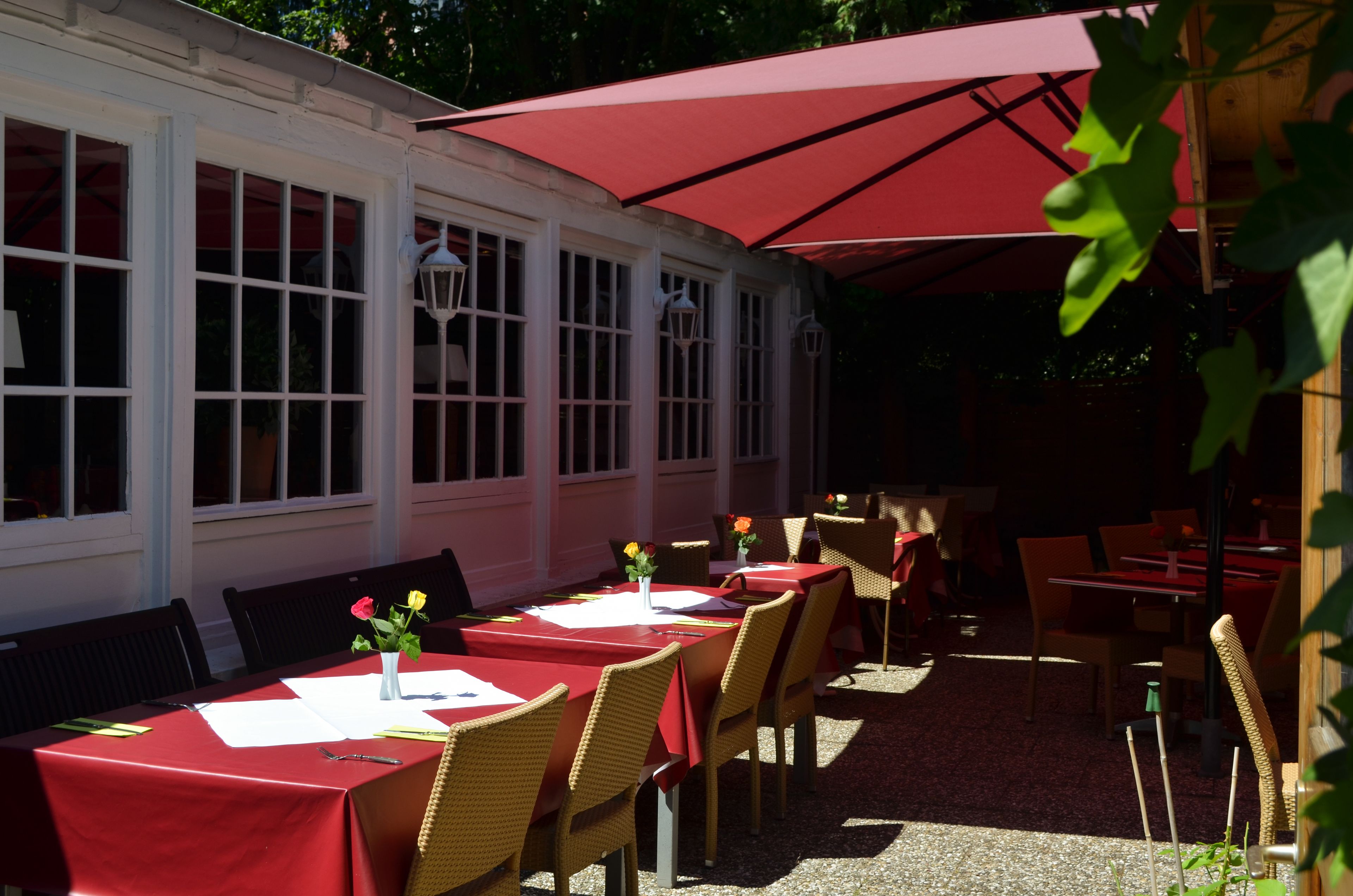 An outdoor dining area with red tables and wicker chairs, covered by a large red umbrella.