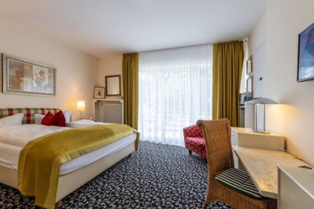 A hotel room with a bed, a desk, a chair, a radiator, and a window with curtains.