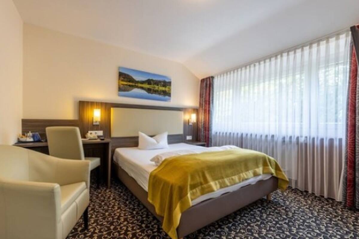 A hotel room with a bed, a desk, a chair, a painting on the wall, and a window with curtains.