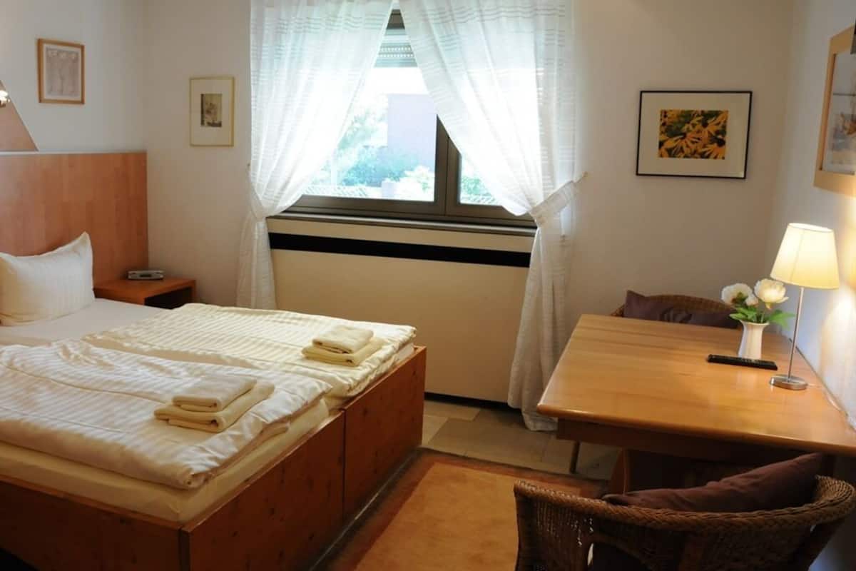 A hotel room with a bed, a wooden table, a chair, a window with curtains, and a lamp.