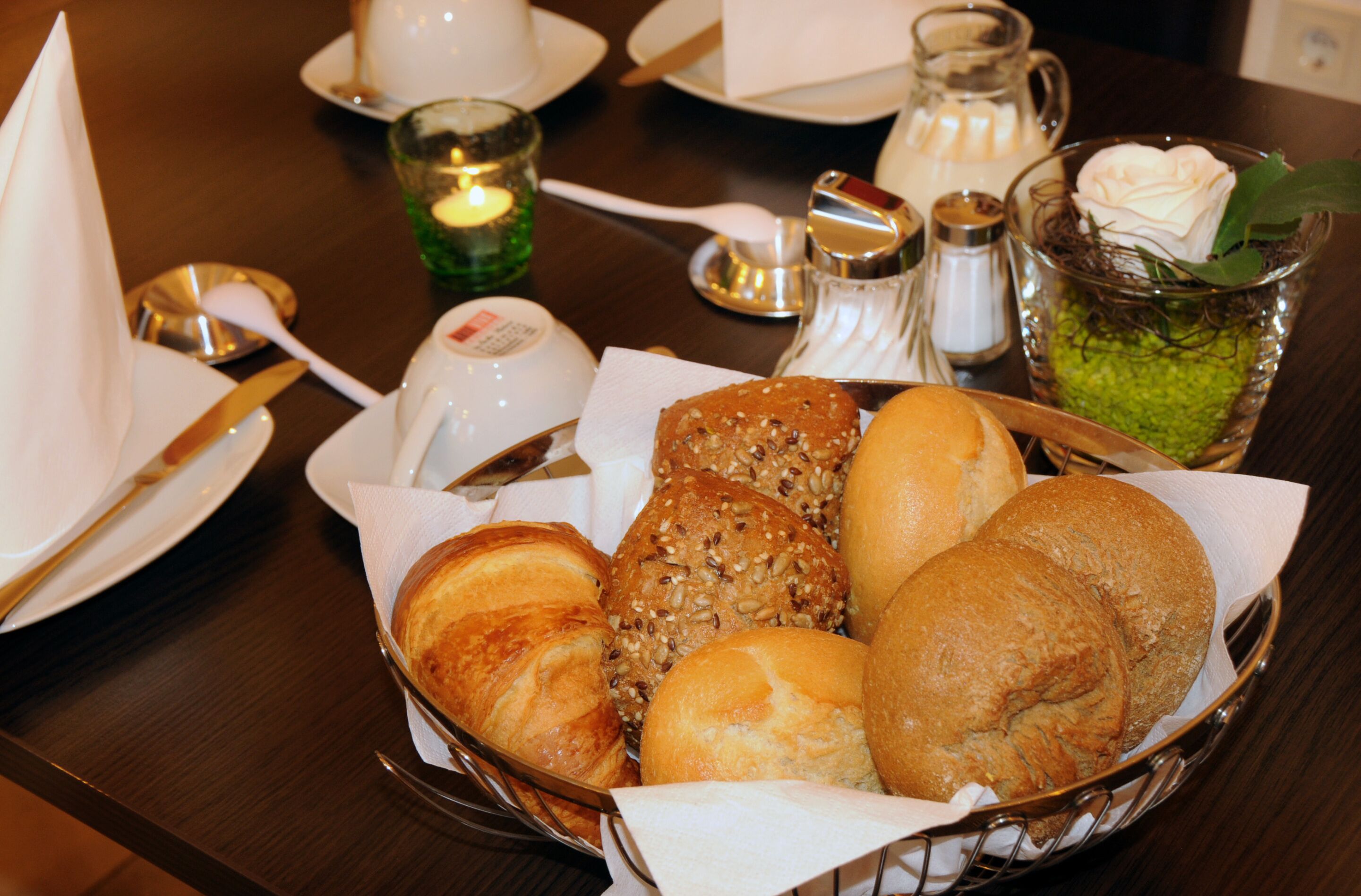 A table with a basket of assorted breads, a candle, salt and pepper shakers, a sugar bowl, and a glass with a rose.