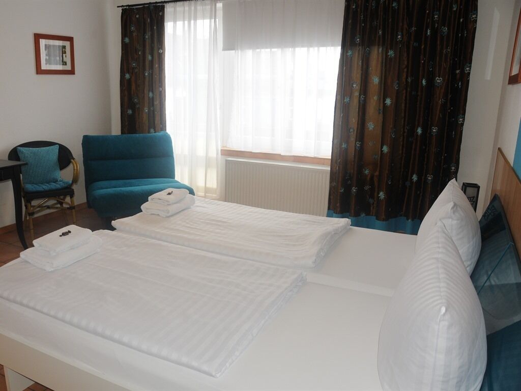 Une chambre d’hôtel avec deux lits, un fauteuil bleu, une petite table et une fenêtre avec des rideaux.