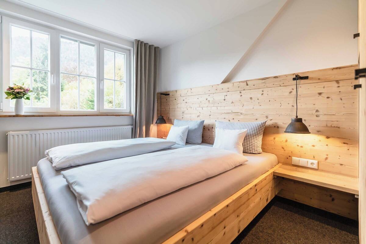 A bedroom with a wooden headboard, a large window with curtains, a bedside table with a lamp, and a radiator.