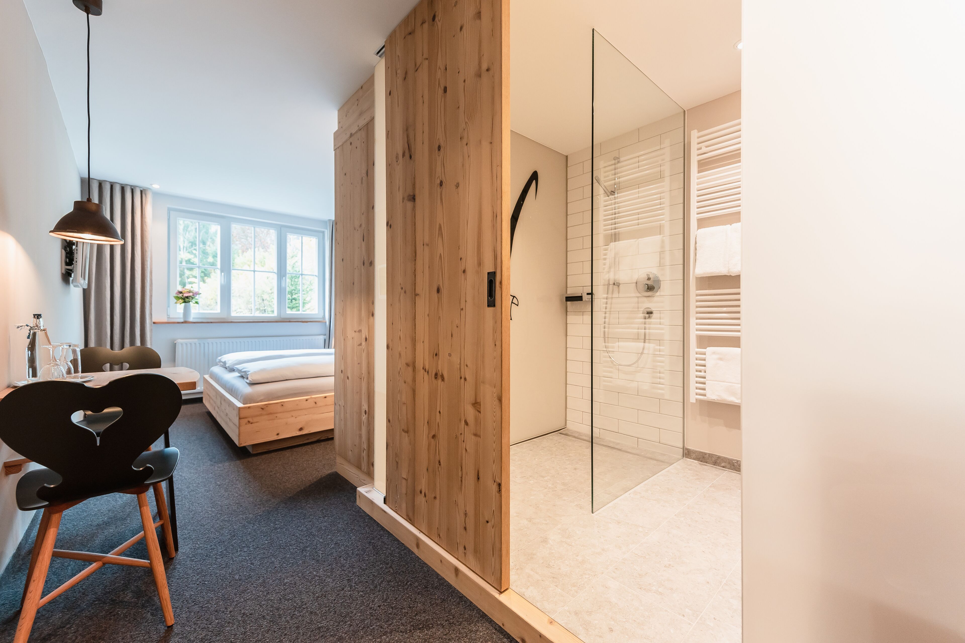 A modern hotel room with a wooden wardrobe, a glass shower enclosure, a bed with a white headboard, and a desk with a chair.