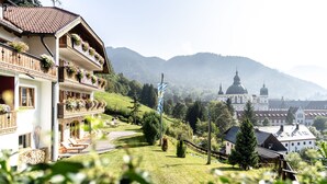 Un hôtel doté de balcons ornés de fleurs, d’un jardin avec des chaises longues et offrant une vue sur une église et des montagnes en arrière-plan.