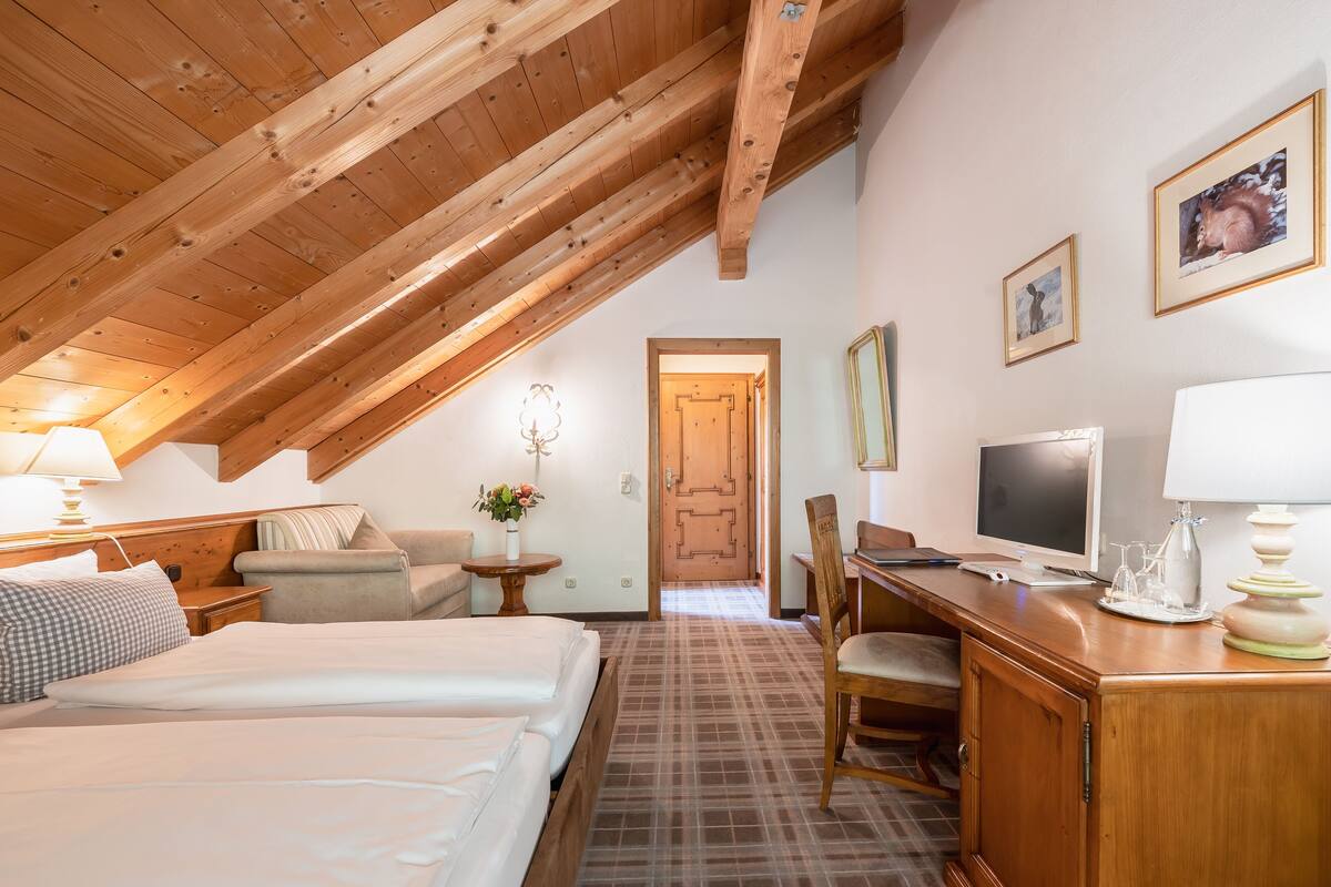 A hotel room with a wooden ceiling, a bed, a desk, a chair, a TV, and a door leading to another room.