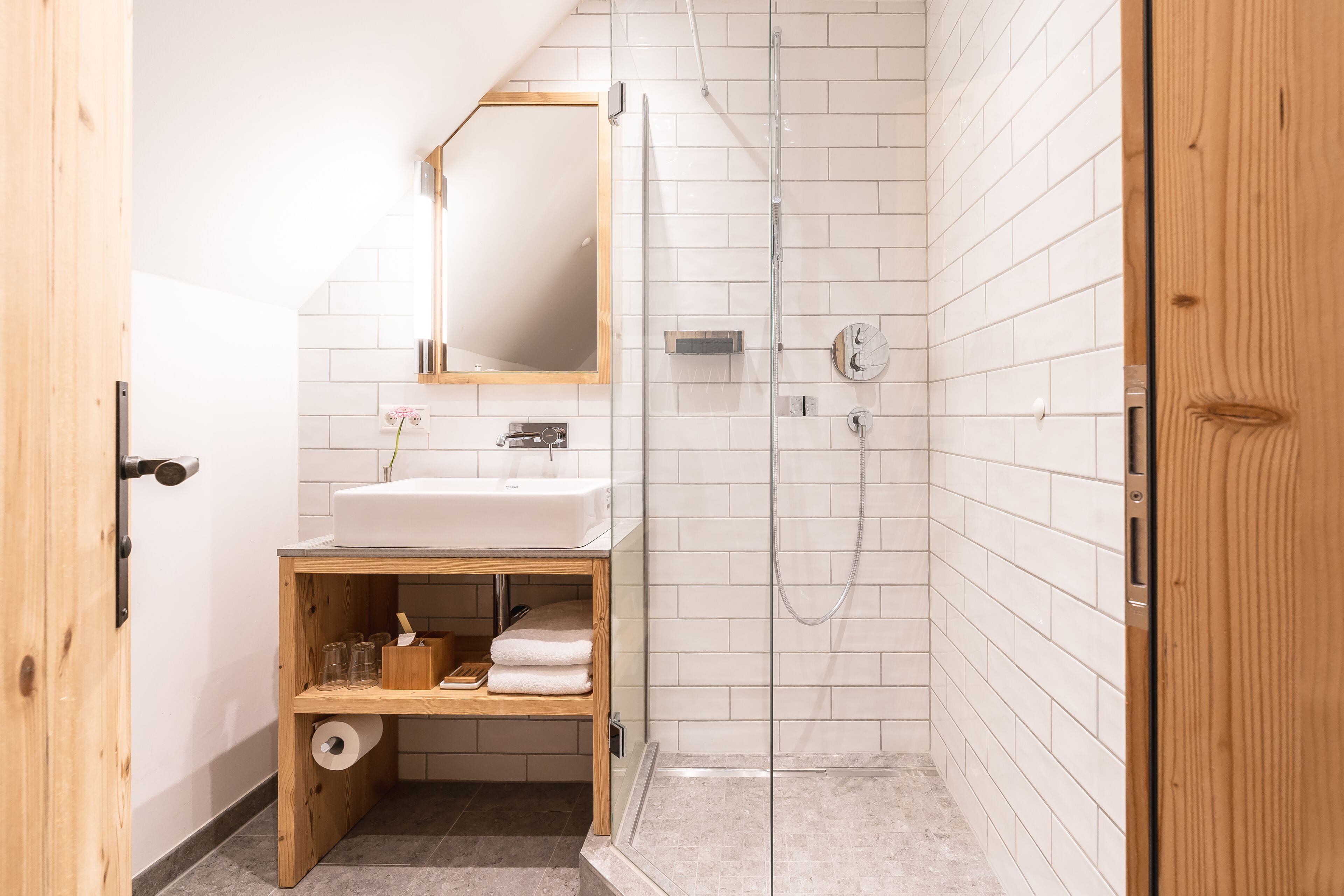 A modern bathroom with a wooden vanity, a glass shower enclosure, and white tiled walls.