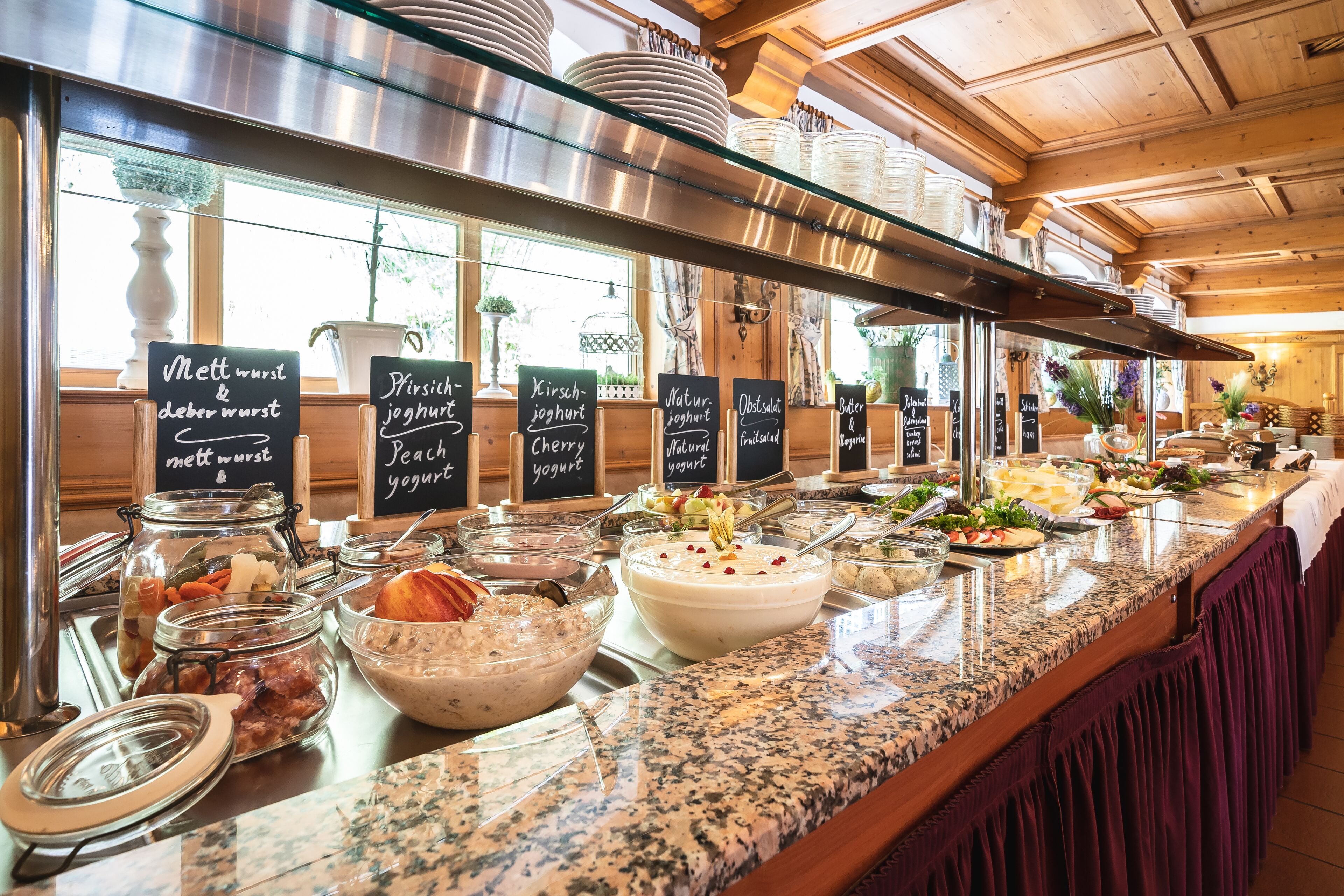 Ein reichhaltiges Buffet mit verschiedenen Speisen und Beschriftungen in einem hellen, holzgetäfelten Innenraum.