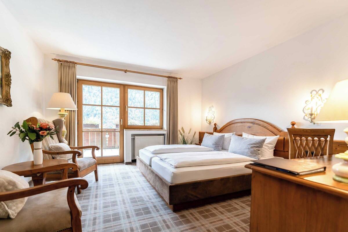 A bedroom with a bed, two chairs, a desk, a window with curtains, and a radiator.