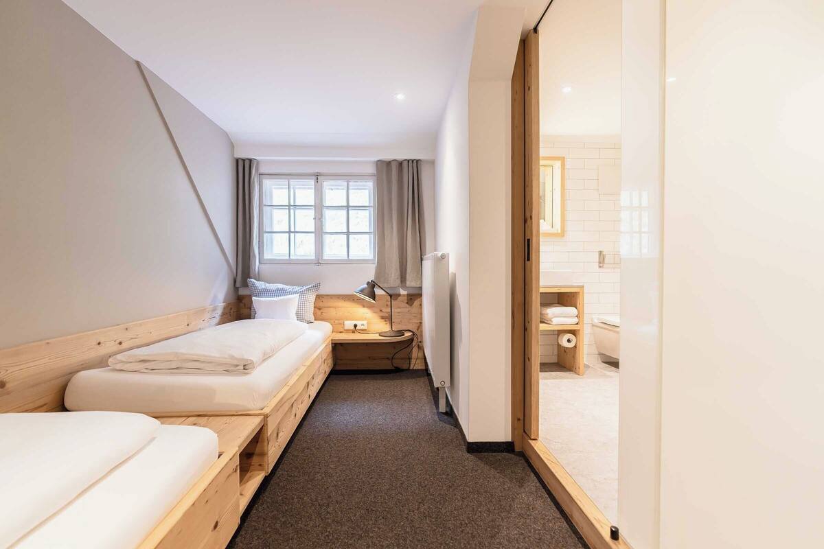 A narrow hotel room with two beds, a small desk, a window with curtains, and a bathroom visible through an open door.