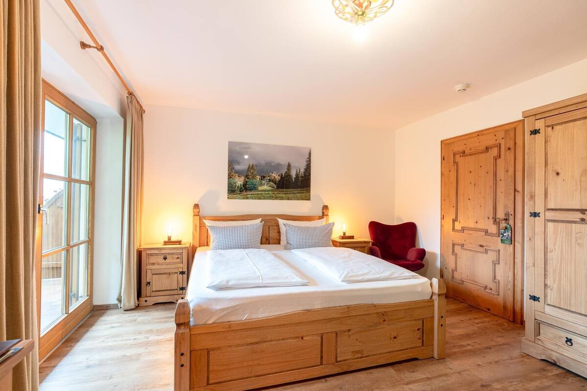 A wooden bed with white linens, a nightstand with a lamp, a red chair, and a large wardrobe.