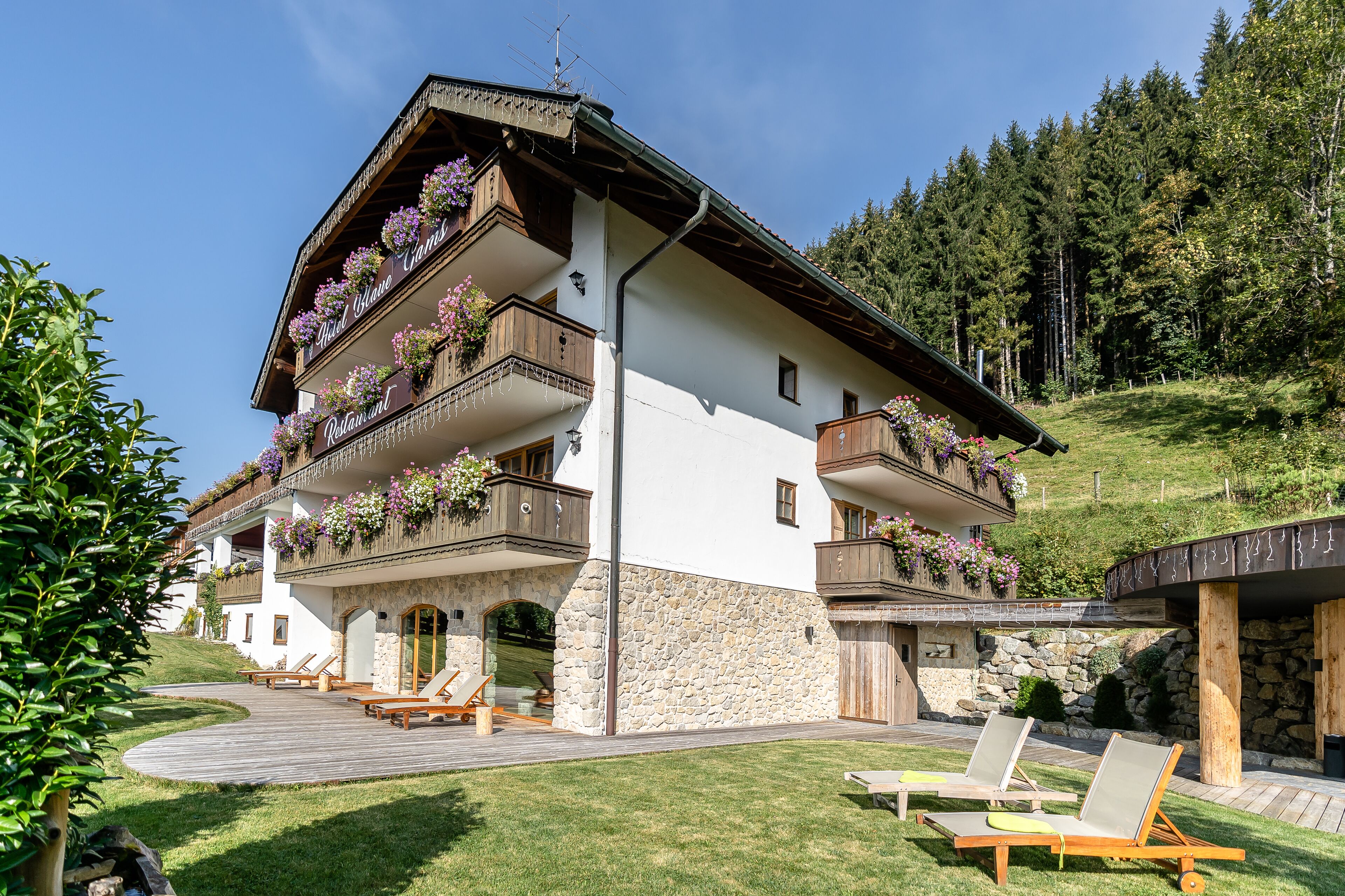A two-story building with balconies adorned with flowers, a stone facade, and a wooden deck with lounge chairs.