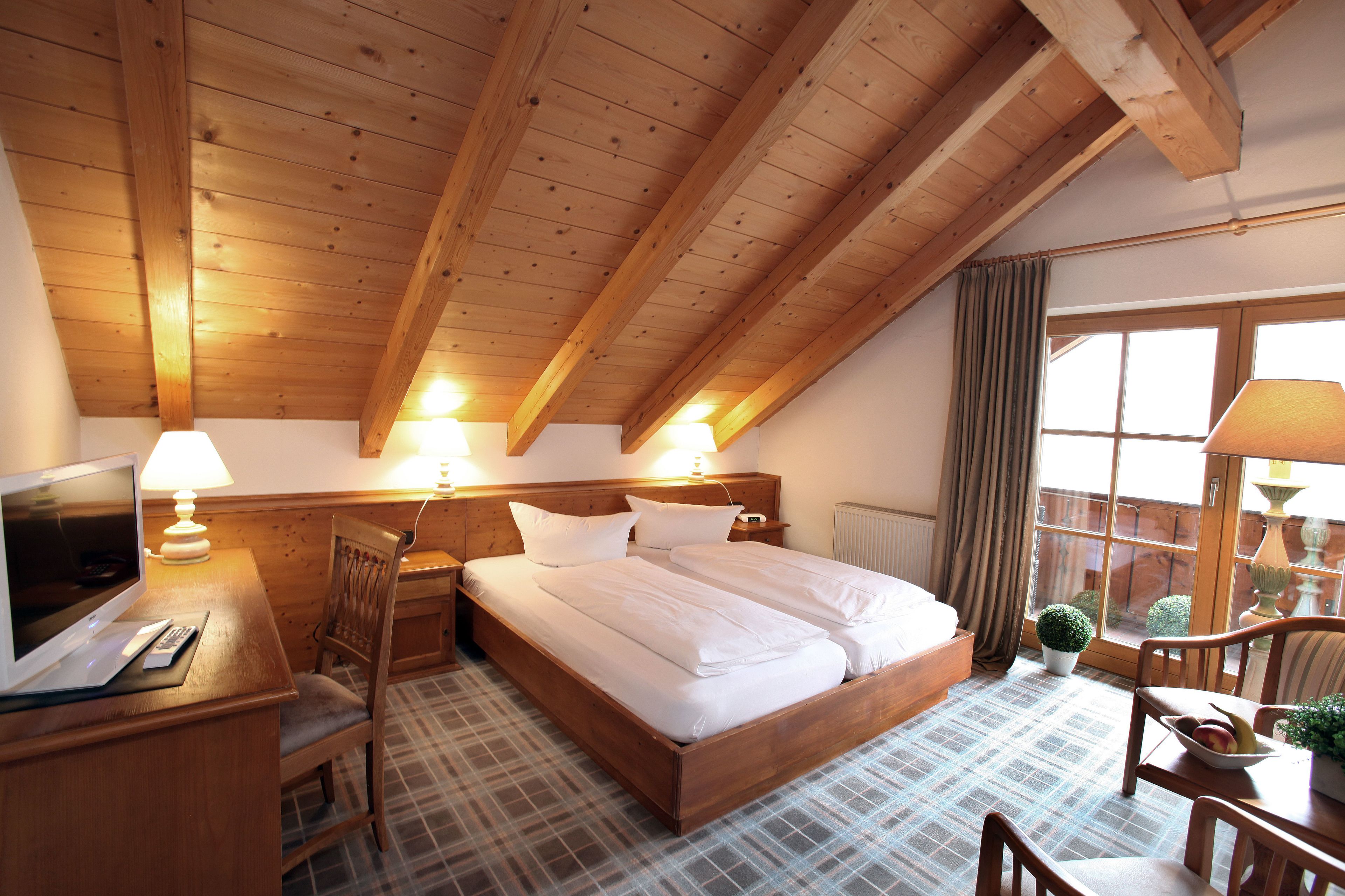 Un dormitorio con techo de madera, una cama, un escritorio con ordenador, una silla y una ventana con cortinas.