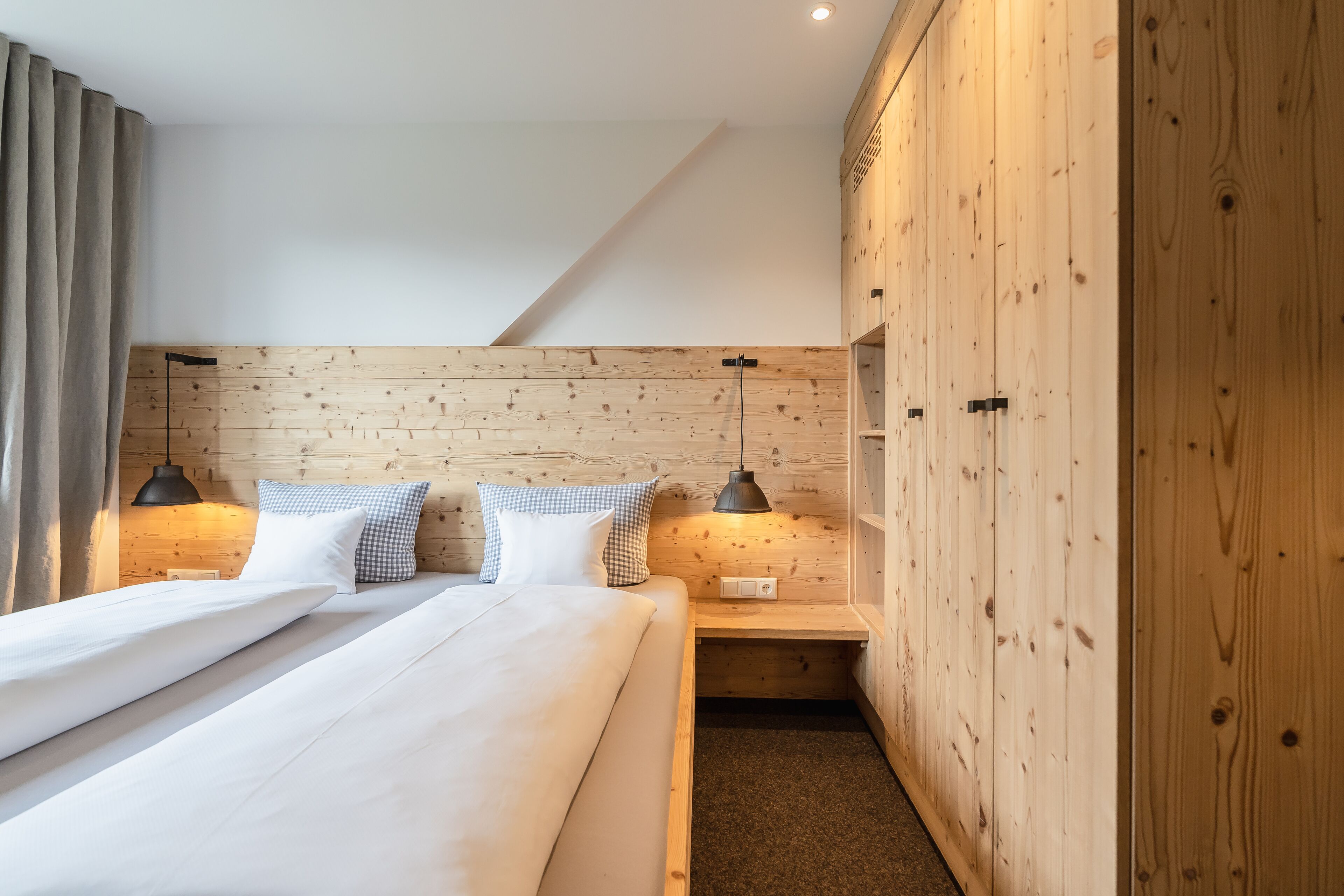 Quarto de hotel moderno com cabeceira de madeira, cama bem arrumada com roupa de cama branca, duas mesas de cabeceira com abajures e um guarda-roupa de madeira.