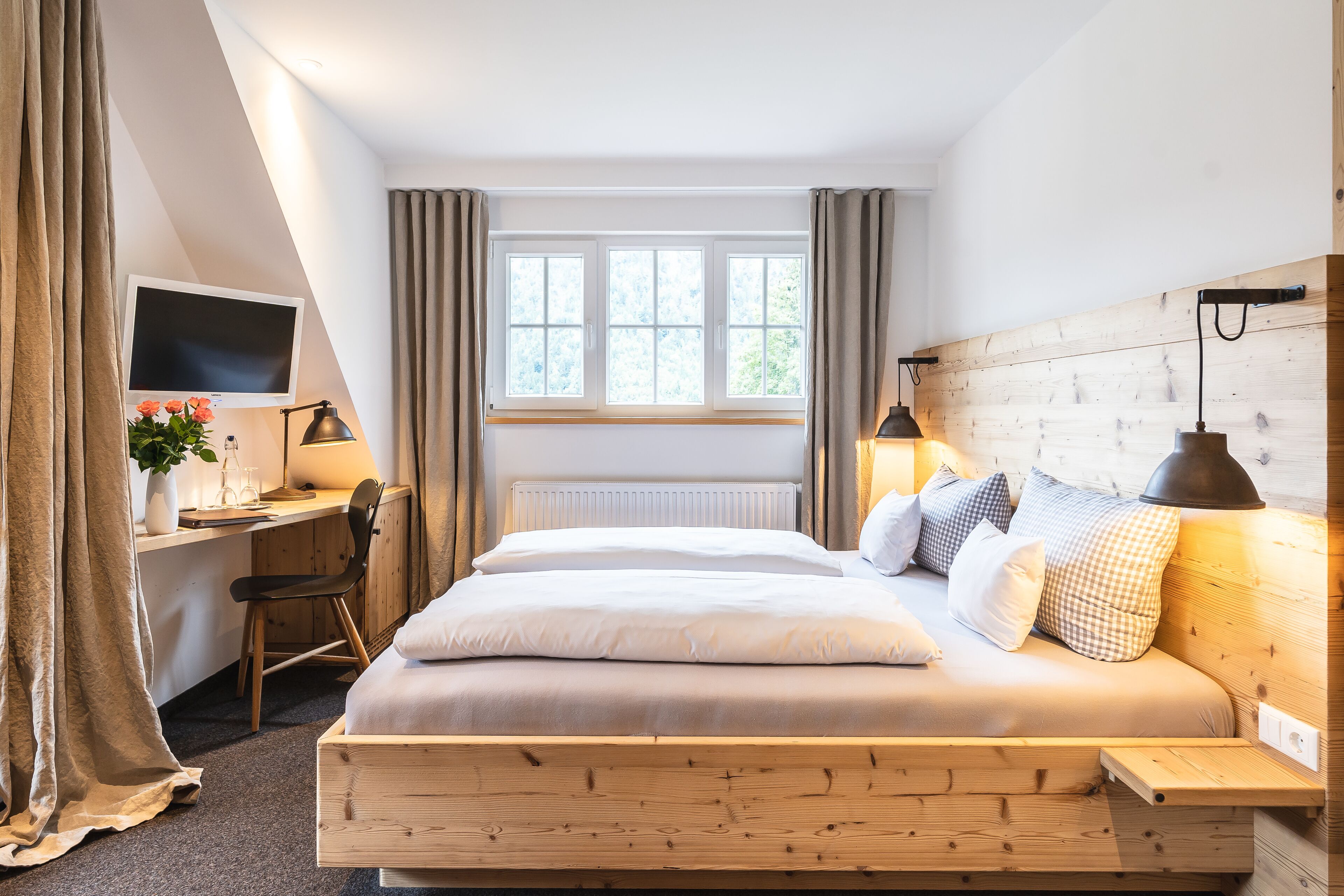 A bedroom with a wooden bed, a desk with a computer, a TV, and a window with curtains.