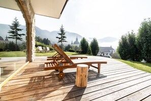 Um deck de madeira com cadeiras de descanso e uma mesa, com vista para uma paisagem montanhosa com uma igreja.