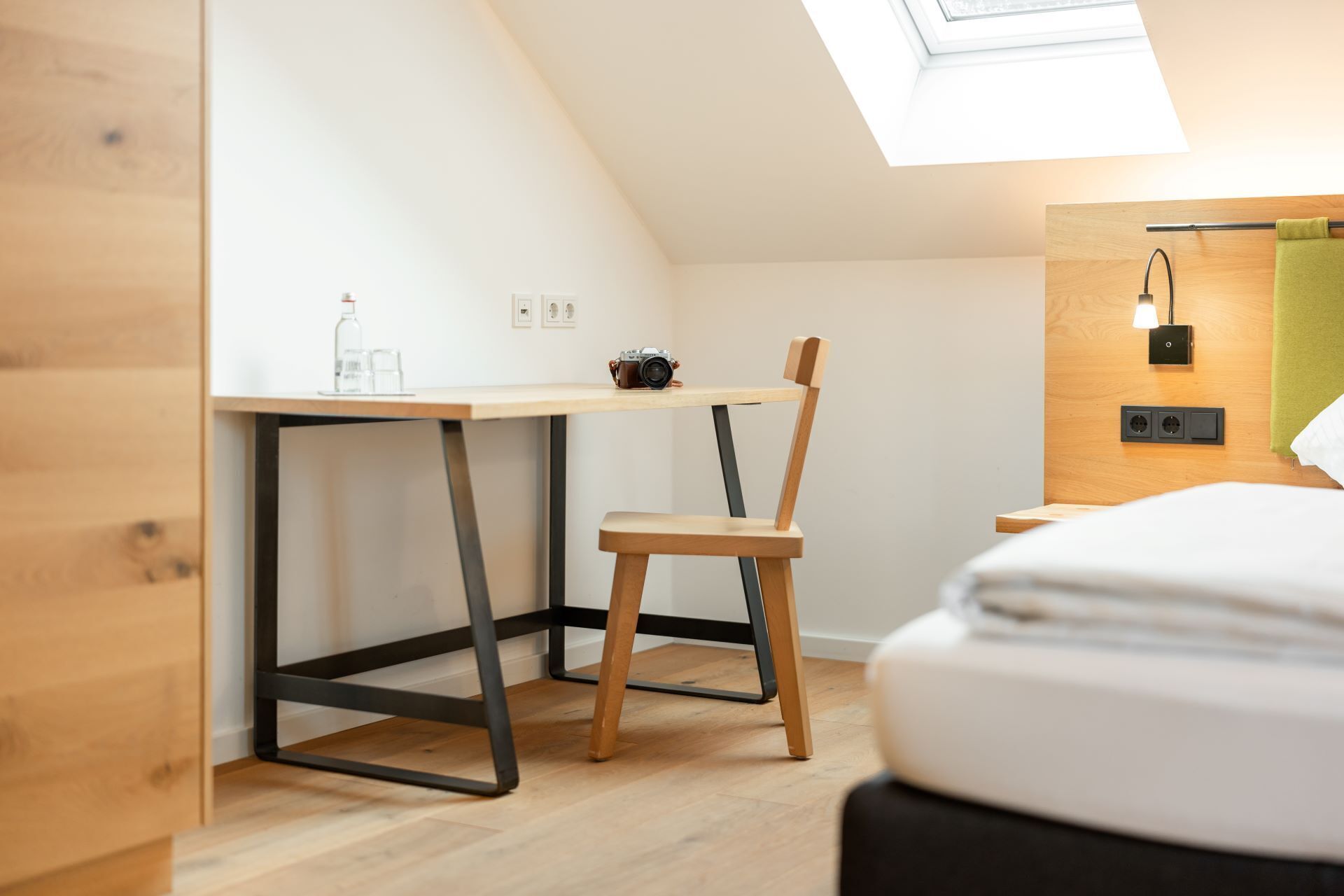 Una habitación moderna y compacta con techo inclinado, un escritorio de madera con una cámara, una silla y una cama con una toalla verde.