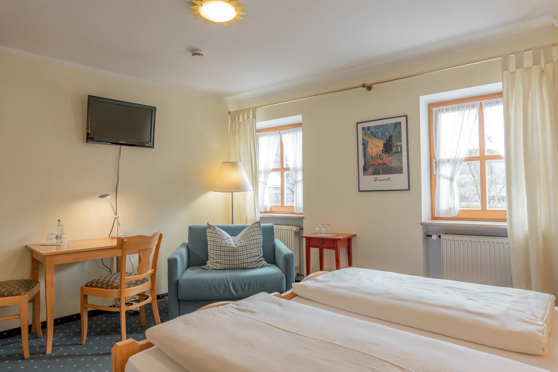 A hotel room with a bed, a desk with a chair, a television on the wall, and two windows with curtains.