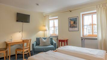A hotel room with a bed, a desk with a chair, a television on the wall, and two windows with curtains.