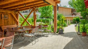 Een terras buiten met houten tafels en stoelen, omgeven door groen en een pad.