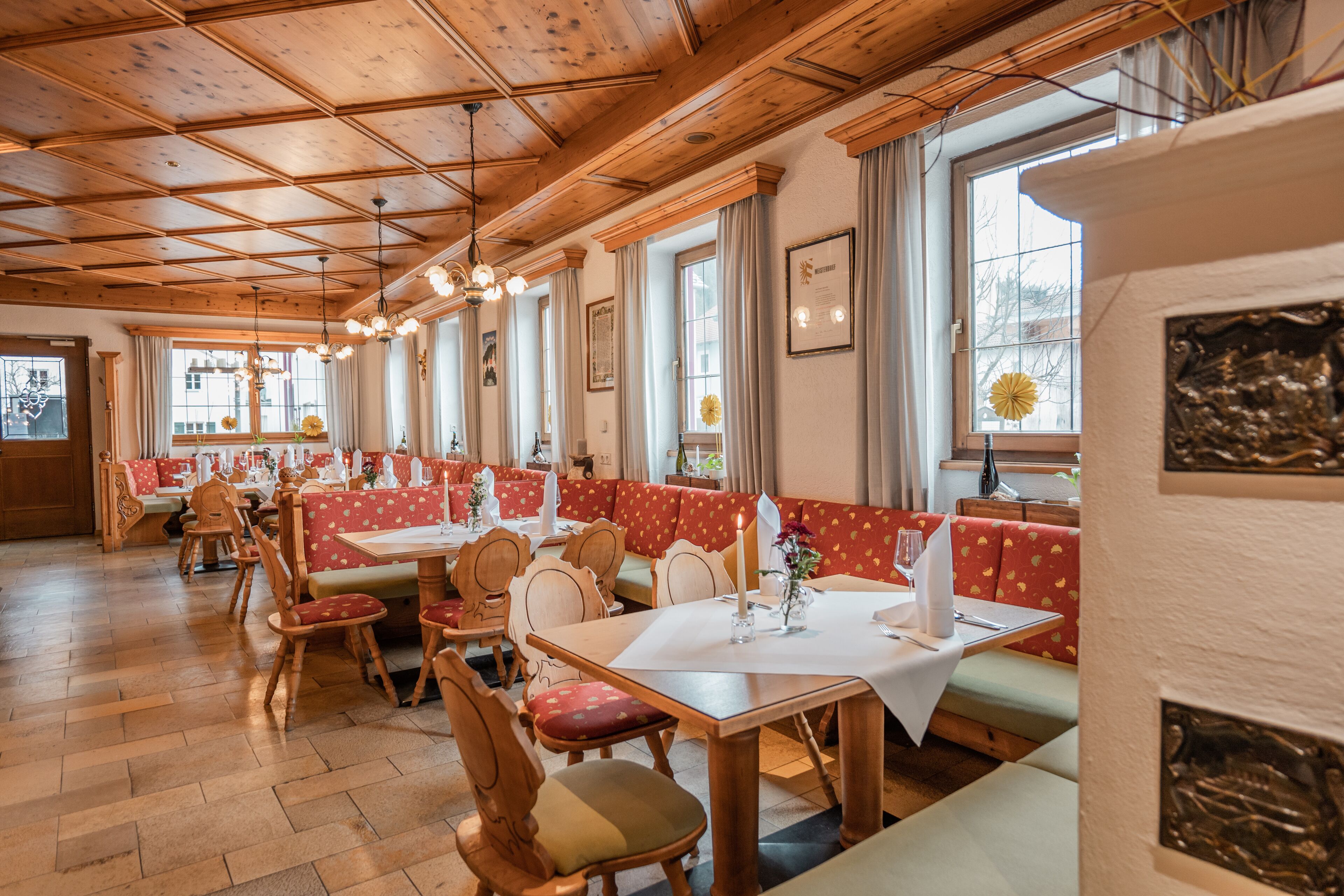 Ein Restaurant mit Holzbalkendecke, rot-weißen Sitzbänken und großen Fenstern mit Vorhängen.