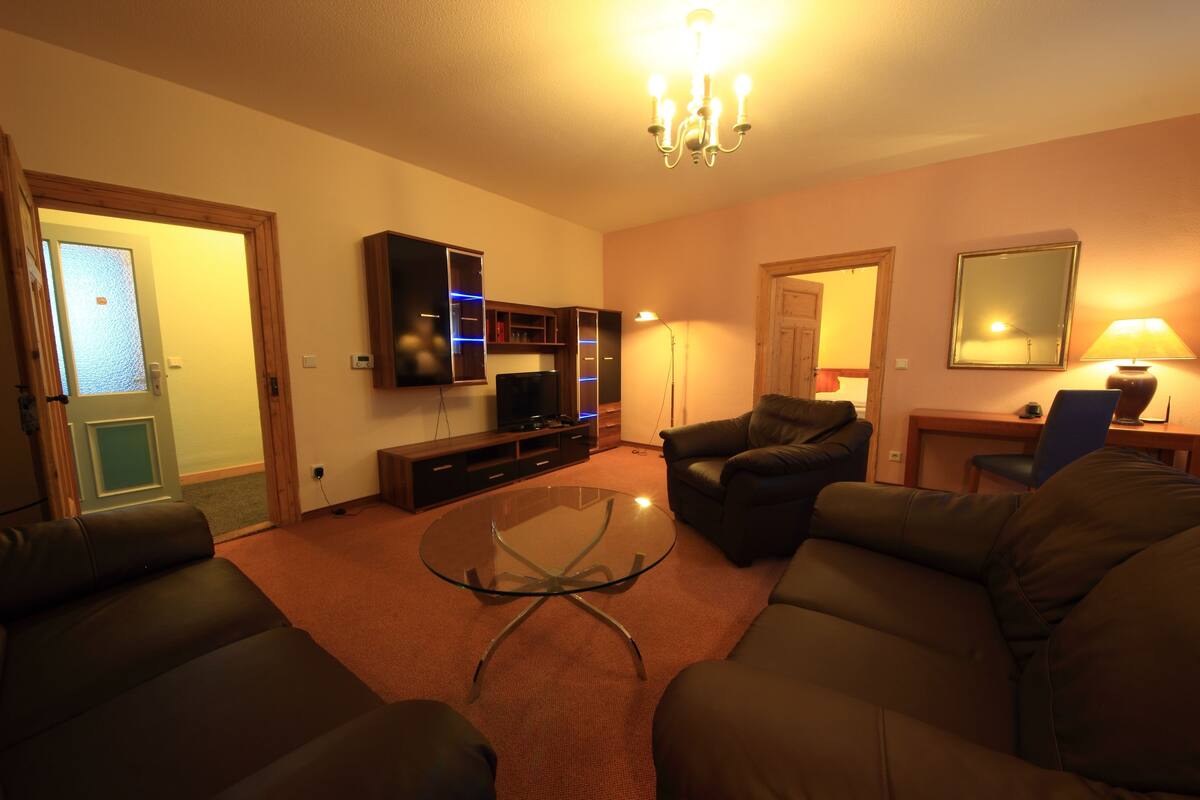 A living room with a glass coffee table, leather sofas, a TV, and a chandelier.