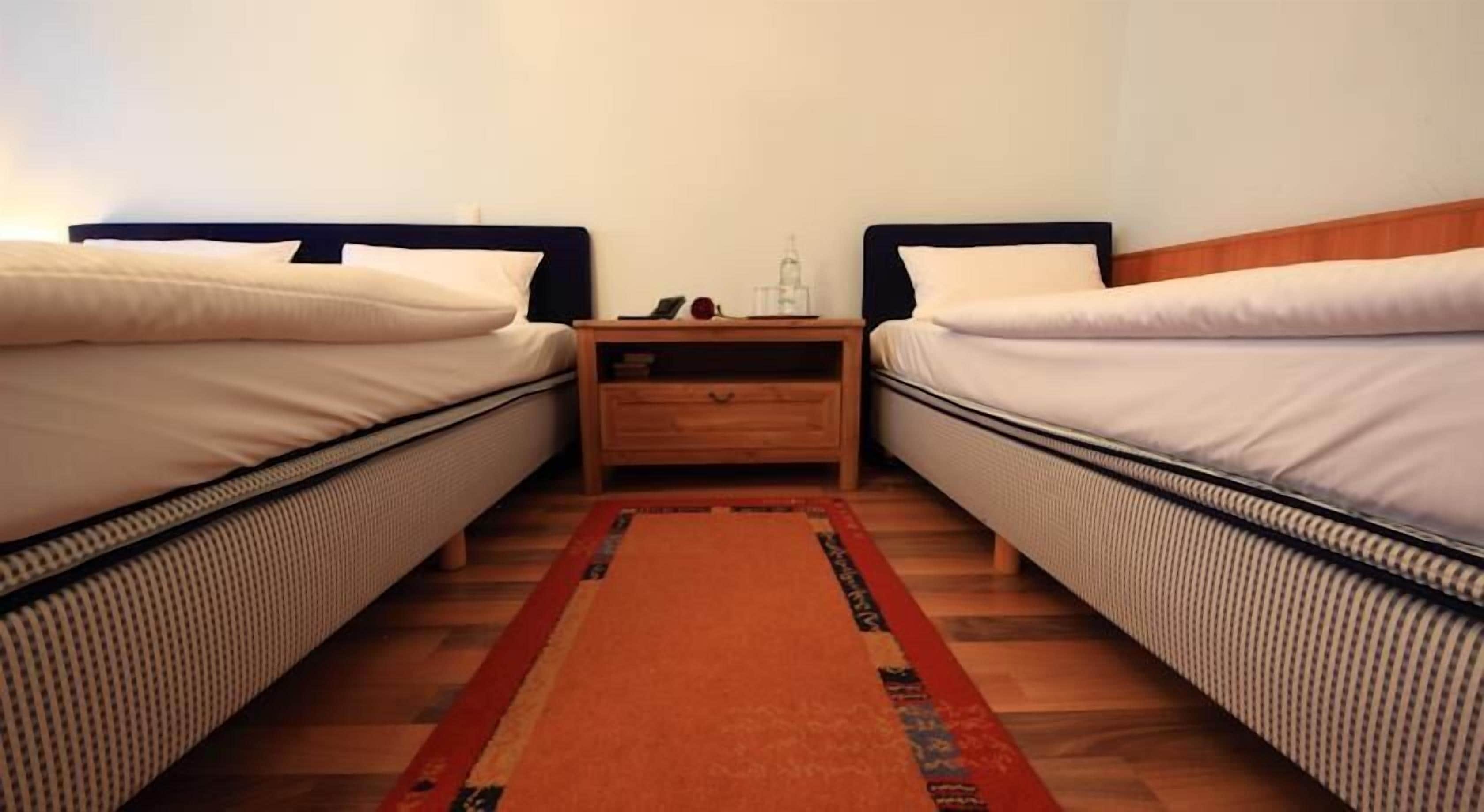 A room with two single beds, a wooden nightstand between them, and a patterned rug on the wooden floor.