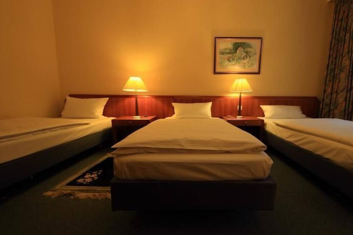 A hotel room with two single beds, a wooden headboard, a painting on the wall, and a window with curtains.