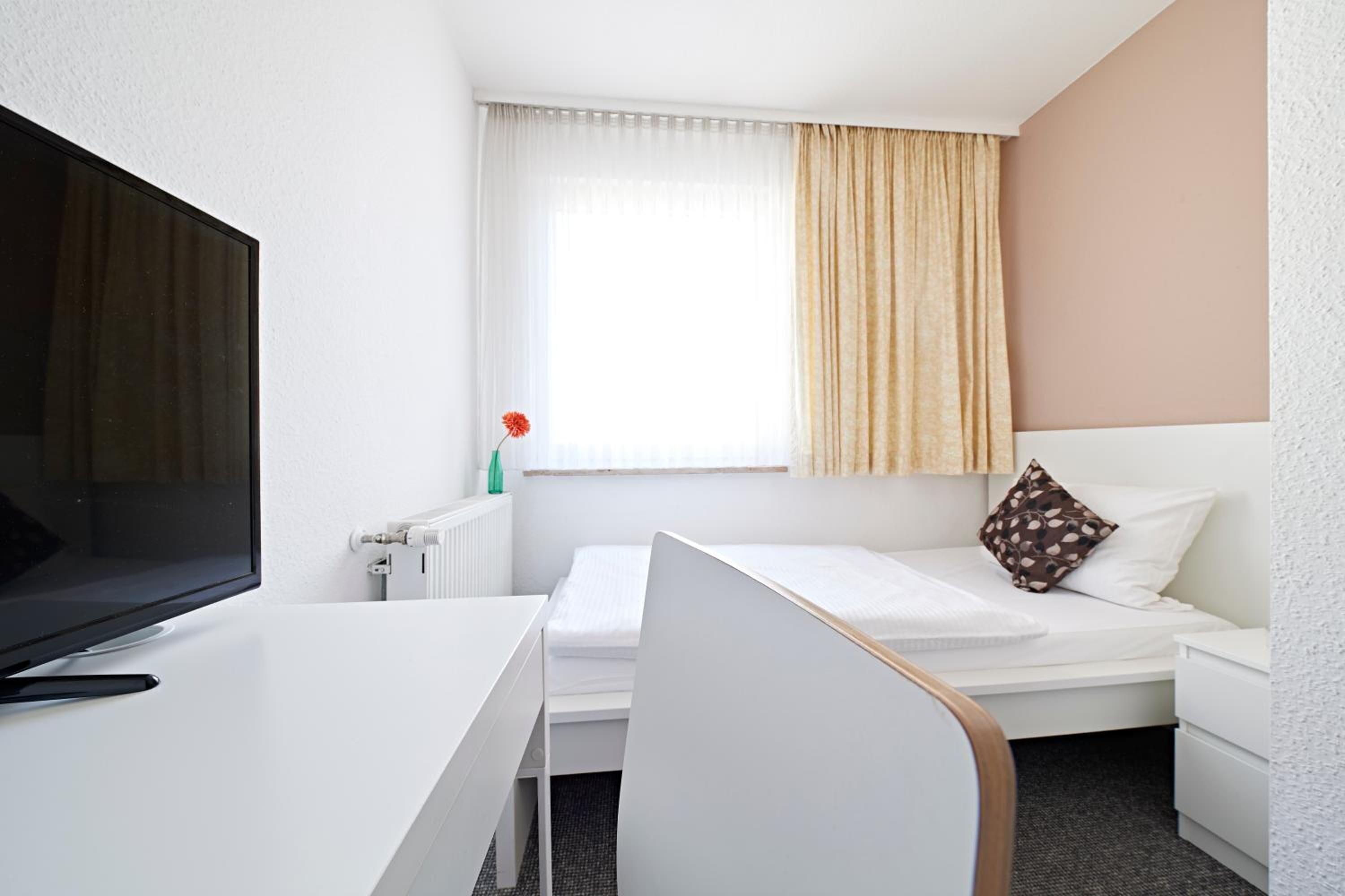 Una camera d'albergo con un letto, una scrivania con la televisione, una finestra con tende e un termosifone.